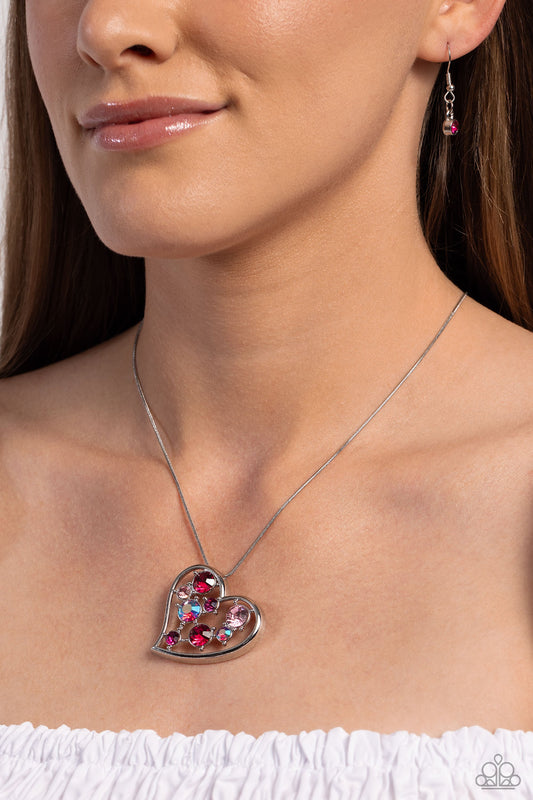 Romantic Recognition - Pink Heart Necklace - Paparazzi Accessories - Cascading from a silver snake chain, a thick, sideways sliding-heart silhouette falls down the neckline for a romantic statement. Haphazardly scattered across the inside of the heart frame, a glittery collection of pink and iridescent fuchsia round rhinestones in varying sizes shimmer and shine for a refined finish.