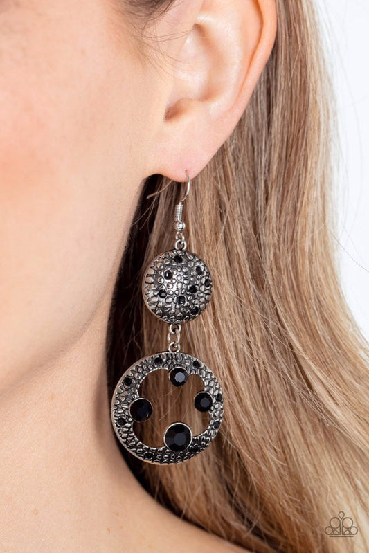 Eastern Entrada - Black and Silver Earrings - Paparazzi Accessories - Dotted with dainty black rhinestones and embossed in pebble-like textures, a beveled silver circle attaches to an imperfect silver hoop featuring matching texture. A smattering of oversized black rhinestones haphazardly adorns the hoop, resulting in a gritty shimmer.