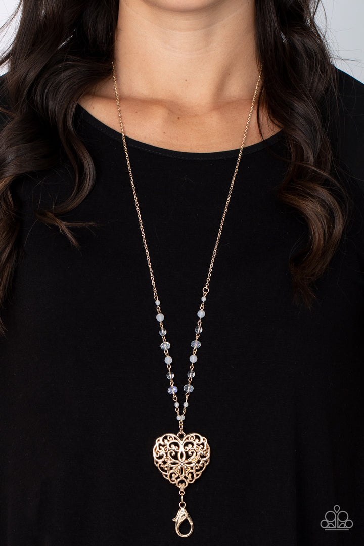 Doting Devotion - Gold Heart Lanyard - Paparazzi Accessories Lanyard - A dainty collection of opaque, glassy, and crystal-like beads give way to an oversized filigree filled heart frame, creating an antiqued locket inspired pendant at the bottom of a lengthened gold chain. A lobster clasp hangs from the bottom of the design to allow a name badge or other item to be attached.