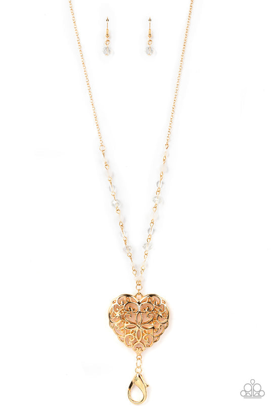 Doting Devotion - Gold Heart Lanyard - Paparazzi Accessories Lanyard - A dainty collection of opaque, glassy, and crystal-like beads give way to an oversized filigree filled heart frame, creating an antiqued locket inspired pendant at the bottom of a lengthened gold chain. A lobster clasp hangs from the bottom of the design to allow a name badge or other item to be attached.